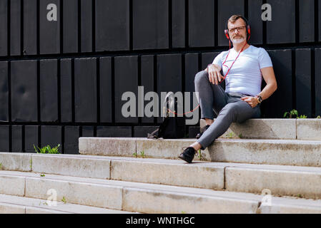 Barbuto uomo maturo seduto sui gradini con le cuffie durante i momenti di relax in una giornata di sole Foto Stock