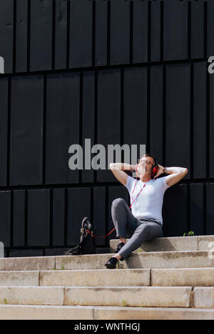 Barbuto uomo maturo seduto sui gradini con le cuffie durante i momenti di relax in una giornata di sole Foto Stock
