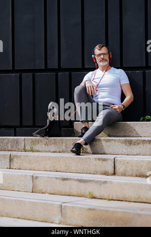 Barbuto uomo maturo seduto sui gradini con le cuffie durante i momenti di relax in una giornata di sole Foto Stock