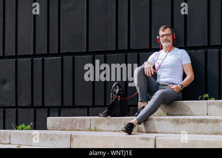 Barbuto uomo maturo seduto sui gradini con le cuffie durante i momenti di relax in una giornata di sole Foto Stock