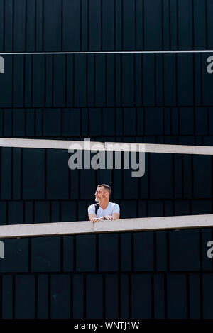 Riflessivo barbuto uomo maturo in t-shirt appoggiata sulla recinzione e denocciolate guardando lontano contro la parete del modello Foto Stock