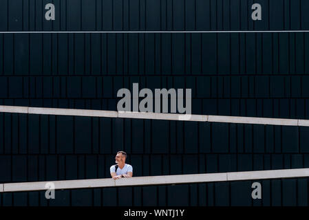 Riflessivo barbuto uomo maturo in t-shirt appoggiata sulla recinzione e denocciolate guardando lontano contro la parete del modello Foto Stock