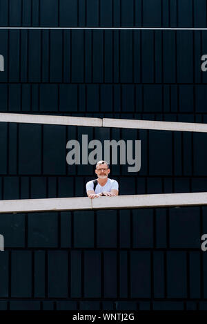 Riflessivo barbuto uomo maturo in t-shirt appoggiata sulla recinzione e denocciolate guardando la telecamera contro la parete del modello Foto Stock