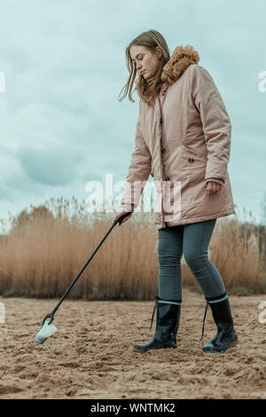 Una giovane donna in stivali di gomma raccoglie i rifiuti in plastica su una spiaggia con una pinza Foto Stock