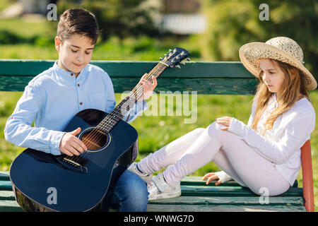 Giovane ragazza e ragazzo con la chitarra seduto al banco nel parco. I bambini amano. Foto Stock