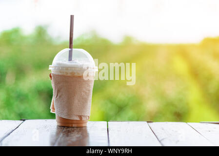 Un take away bicchiere di iced cappuccino caffè è sul tavolo di legno con sfondo sfocato verde natura outdoor tra di sole in estate mo Foto Stock