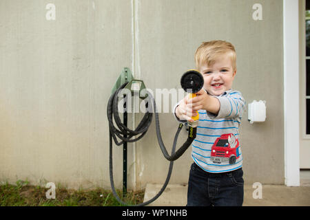 Giocoso adorabile toddler boy guarda avanti che trattengono il tubo flessibile a spruzzo per spruzzare in avanti Foto Stock