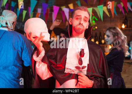 Attraente uomo vestito come vampire con un teschio umano alla festa di Halloween. Creepy vampiro alla festa di Halloween. Foto Stock
