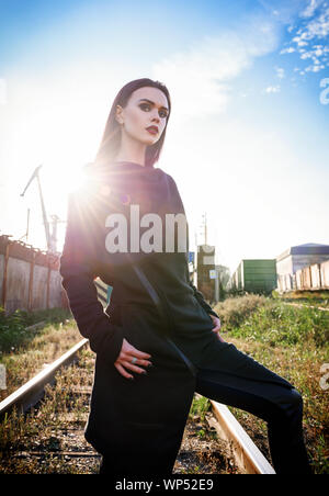 Moda shot: ritratto della bella ragazza rock (modello informale) in tunica e pantaloni di pelle in piedi alla ferrovia (zona industriale) Foto Stock