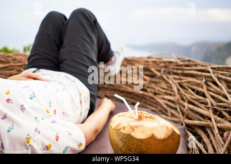 Giovane donna prendendo una pausa e bere una noce di cocco dentro un nido di fronte al mare, Bali, Indonesia Foto Stock