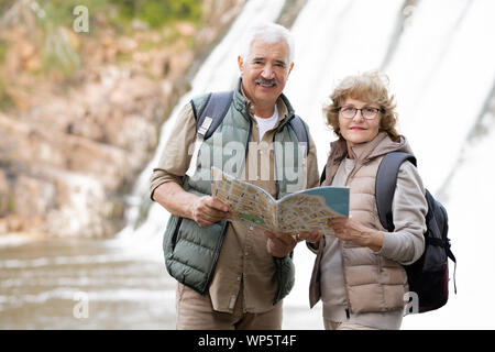 Felice coppia senior con zaini e mappa in piedi nella parte anteriore della fotocamera Foto Stock