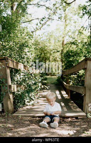 Piccolo grazioso dai capelli biondi Toddler Boy Kid bambino seduto e ridere nella parte anteriore del ponte di legno su un torrente presso il parco nel bosco durante la stagione estiva