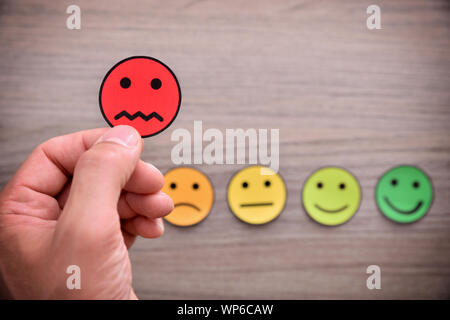 Mano tenendo una valutazione negativa rappresentata con ritagli di faccia con espressioni diverse sul tavolo di legno. Composizione orizzontale. Vista dall'alto. Foto Stock