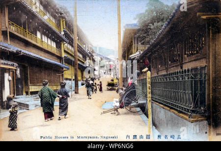 [ 1910s Giappone - bordelli a Nagasaki il distretto a luci rosse ] - La prostituzione distretto di Maruyama (丸山町) in Nagasaki. Un jinrikisha (rickshaw) estrattore si siede nel suo rickshaw, le donne e i bambini a piedi sulla strada fiancheggiata con tre storia bordelli. Xx secolo cartolina vintage. Foto Stock