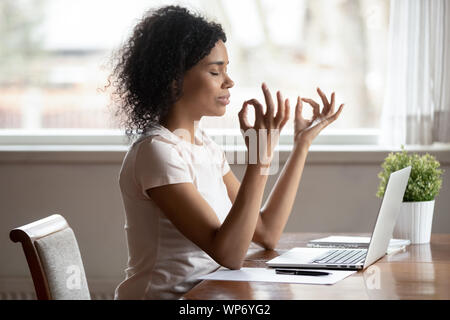 Calma biracial donna meditare vicino al computer portatile al lavoro Foto Stock