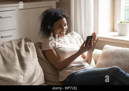 Felice biracial donna nelle cuffie utilizza lo smartphone a casa Foto Stock