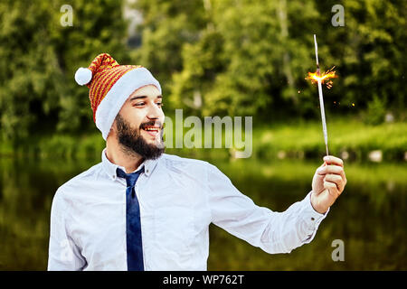 Un imprenditore in un divertente Santa Claus hat guarda un ardente sparkler nella sua mano. Il suo ufficio festeggia il nuovo anno, o il Natale. Un sparkler brucia in th Foto Stock