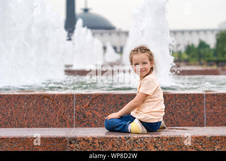 Carino piccolo maliziosa bambina gioca presso la fontana. Tempo della beffa. Urbano abbigliamento casual. Spensierato. La felicità, divertente e concetto di infanzia. Foto Stock