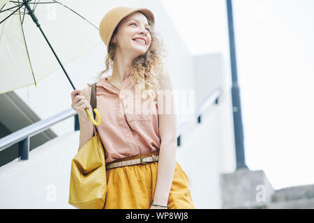 Attraente felice giovane donna in hat passeggiate all'aperto con ombrello sul giorno di pioggia Foto Stock