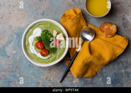 Zuppa di broccoli nella ciotola con il prezzemolo e i pomodori, con cucchiaio e tovagliolo su cemento. Vista superiore, laici piatta Foto Stock