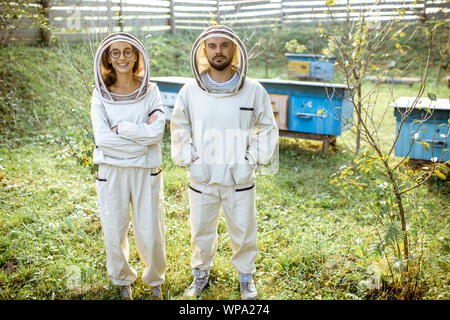 Ritratto di un uomo e di una donna apiarists in uniforme di protezione in piedi su un piccolo tradizionale apiario con alveari in legno sullo sfondo Foto Stock