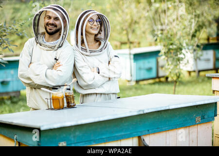 Ritratto di un uomo e di una donna gli apicoltori in uniforme di protezione in piedi insieme sul piccolo tradizionale apiario con alveari in legno Foto Stock