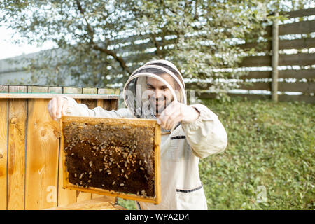 Apicoltore in uniforme di protezione di ottenere nidi d'ape dall'alveare in legno, lavorando sull'apiario Foto Stock