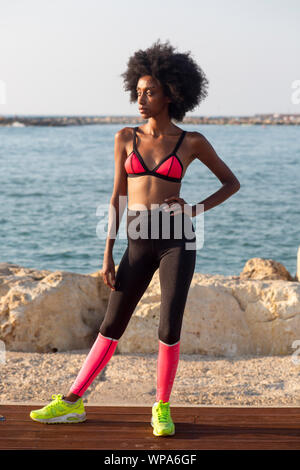 Felice giovane donna africana di stretching e riscaldamento prima di un allenamento all'aperto. Il mare Mediterraneo in background Foto Stock