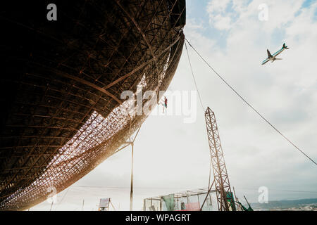 L'uomo scalata di un enorme parabola satellitare durante il tramonto vicino ad un aereo Foto Stock
