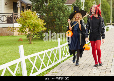 A piena lunghezza ritratto di due piccolo felice le ragazze che indossano costumi di Halloween a piedi in strada mentre il trucco-o-trattare nel quartiere accogliente, spazio di copia Foto Stock