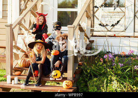 Multi-etnico gruppo di bambini che indossano costumi di Halloween guardando la fotocamera mentre è seduto sulle scale di casa decorata in Trick or Treat stagione, spazio di copia Foto Stock