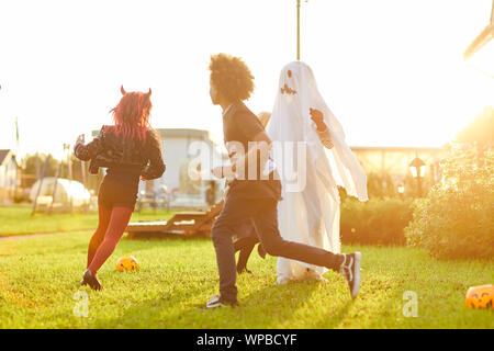 Multi-etnico gruppo di bambini indossando costumi in esecuzione sul prato verde divertendosi su Halloween e i giochi di luce del sole, spazio di copia Foto Stock