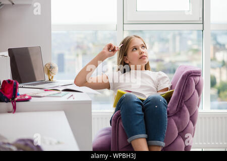 Gioiosa ragazza con una matita nei pressi di capo cercando di pensare a sognare. Forniture scolastiche e laptop in background. Foto Stock