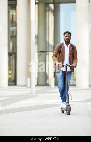 A piena lunghezza Ritratto di contemporaneo afro-uomo che cavalca scooter elettrico e guardando la telecamera durante gli spostamenti nelle strade cittadine, spazio di copia Foto Stock