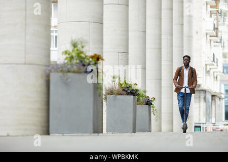 Ampio angolo ritratto di africana contemporanea di equitazione uomo scooter elettrico in città strada andando verso la telecamera, spazio di copia Foto Stock