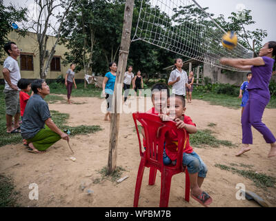 Due giovani bambini vietnamiti seduti sul rosso sedie di plastica con la famiglia e gli amici a giocare a pallavolo in background, Phong Nha, Vietnam Foto Stock