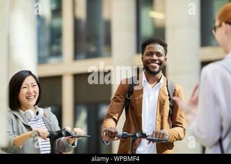 Multi-gruppo etnico di allegro giovani riding scooter elettrici in una strada di città e incontro con gli amici, spazio di copia Foto Stock