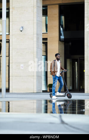 A piena lunghezza Ritratto di contemporaneo afro-uomo che cavalca scooter elettrico nel contesto urbano all'aperto, spazio di copia Foto Stock