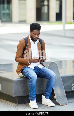 A piena lunghezza Ritratto di giovane uomo afro-americano la digitazione di un messaggio di testo tramite smartphone mentre seduti all'aperto nel contesto urbano, spazio di copia Foto Stock