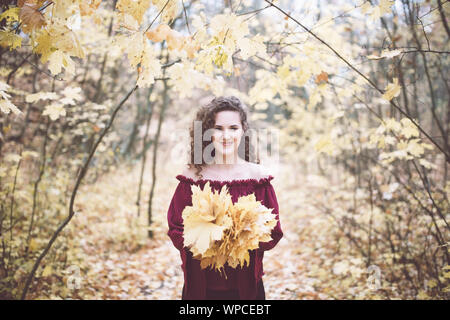 Bellissima ragazza con ricci capelli scuri in un marrone rossiccio top in un autunno park holding foglie di acero e sorridente alla fotocamera Foto Stock