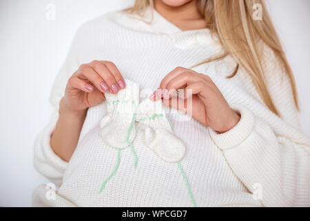 La gravidanza di una ragazza bionda in un maglione bianco tiene nelle sue mani appena lavorato a maglia calze bambino Foto Stock