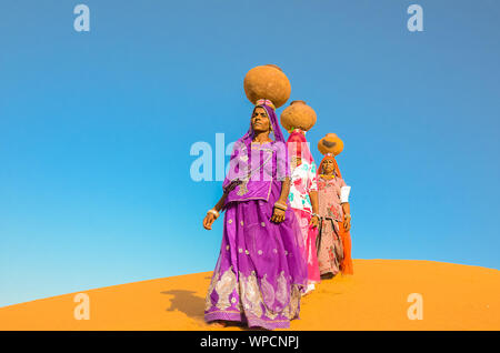 Jaisalmer, Rajasthan, India - aprile 18th, 2018: le donne portano pesanti brocche di acqua sulla loro testa e camminare su un giallo duna di sabbia in estate calda d Foto Stock