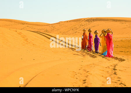 Jaisalmer, Rajasthan, India - aprile 18th, 2018: le donne portano pesanti brocche di acqua sulla loro testa e camminare su un giallo duna di sabbia in estate calda d Foto Stock