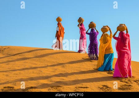 Jaisalmer, Rajasthan, India - aprile 18th, 2018: le donne portano pesanti brocche di acqua sulla loro testa e camminare su un giallo duna di sabbia in estate calda d Foto Stock