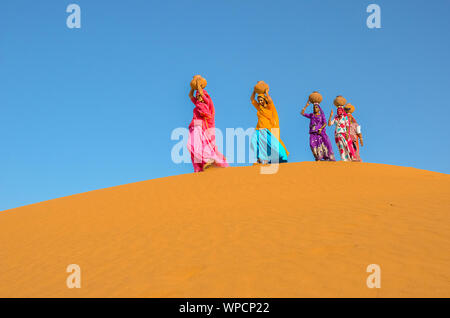 Jaisalmer, Rajasthan, India - aprile 18th, 2018: le donne portano pesanti brocche di acqua sulla loro testa e camminare su un giallo duna di sabbia in estate calda d Foto Stock