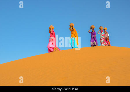 Jaisalmer, Rajasthan, India - aprile 18th, 2018: le donne portano pesanti brocche di acqua sulla loro testa e camminare su un giallo duna di sabbia in estate calda d Foto Stock