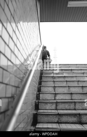 MATSUYAMA, Giappone - Luglio 20, 2019: una verticale in scala di grigi a bassa angolazione di una persona seduta in cima alle scale in prossimità di un corrimano su una parete Foto Stock