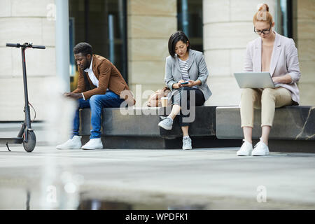 A piena lunghezza ritratto contemporaneo di giovani usando dispositivi elettronici mentre posa sul banco di prova all'aperto nel contesto urbano, spazio di copia Foto Stock