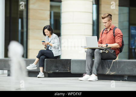 A piena lunghezza ritratto di due giovani usando dispositivi elettronici mentre posa sul banco di prova all'aperto nel contesto urbano, spazio di copia Foto Stock