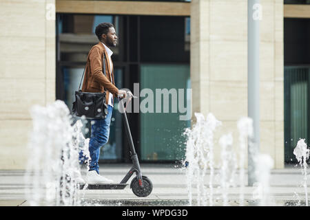 Vista laterale per tutta la lunghezza della moderna afro-americano di uomo che cavalca scooter elettrico attraverso la fontana durante gli spostamenti di lavoro in città, spazio di copia Foto Stock